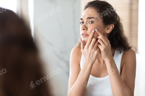 Preview: Unhappy Female Squeezing Pimples On Cheek Looking At Mirror Indoor