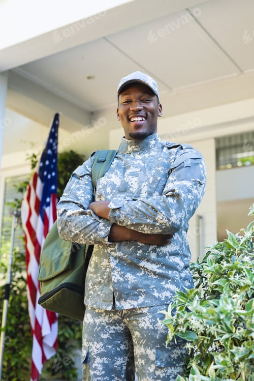 Preview: Happy african american male soldier with arms crossed in cap outside home with usa flag, copy space