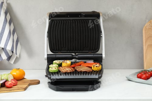Preview: Electric grill with tasty meat and vegetables on white marble table in kitchen