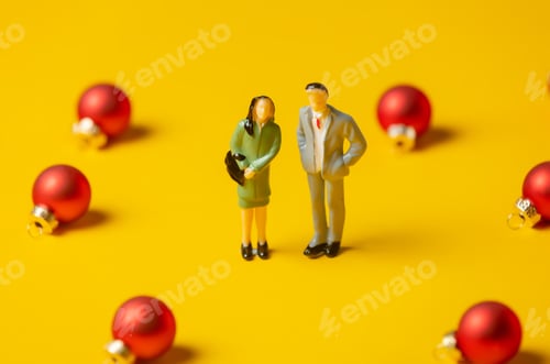 Preview: miniature woman and man figures stand surrounded by red Christmas baubles on yellow background