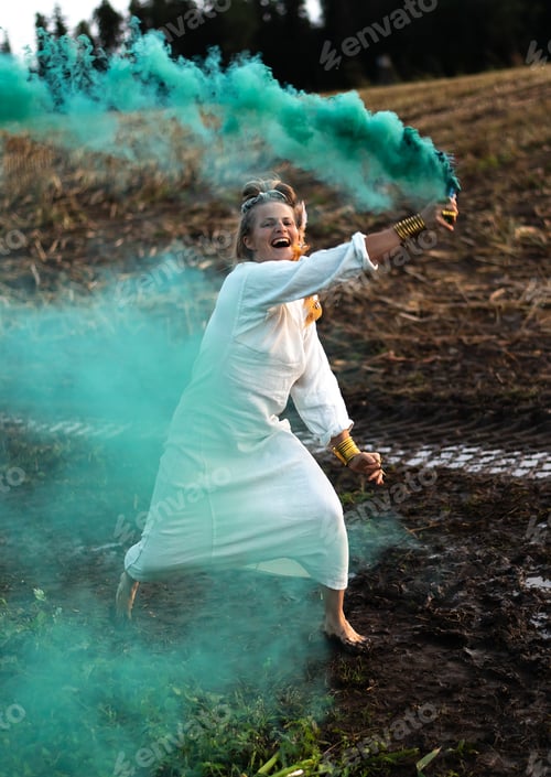 Preview: Happy woman in white dress with green smoke bomb dancing on muddy field