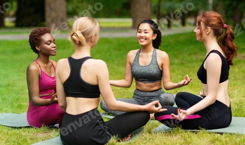 Preview: Diverse young women sitting in circle and practicing yoga meditation at park