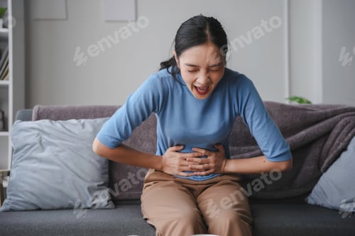 Preview: Young woman experiencing intense stomach pain on sofa at home