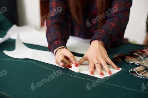 Preview: Tailor worktable in studio. Female hands making chalk markings of paper patterns on green fabric.