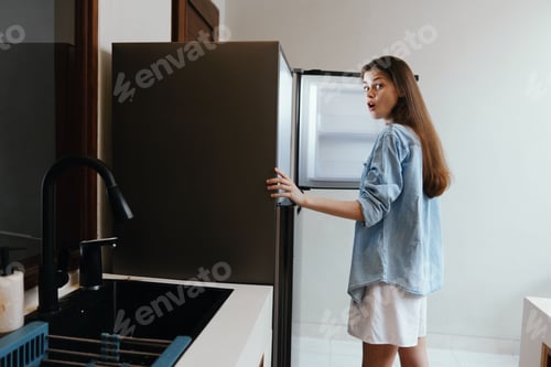 Preview: Surprised young woman discovering an empty fridge in a modern kitchen, showcasing a minimalist
