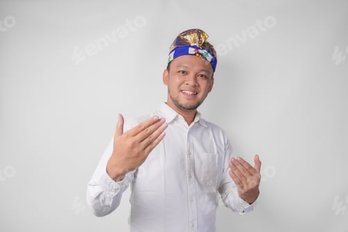 Preview: Young Balinese handsome man in traditional headdress inviting to come with hand.