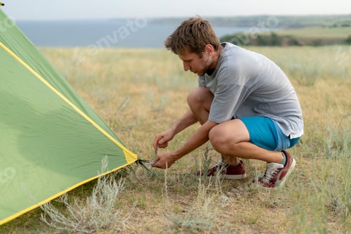Preview: person is kneeling on the ground, securing a tent peg to the ground near a tent.