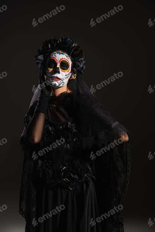 Preview: woman in mexican catrina makeup and dark costume with lace veil posing with hand on hip on black
