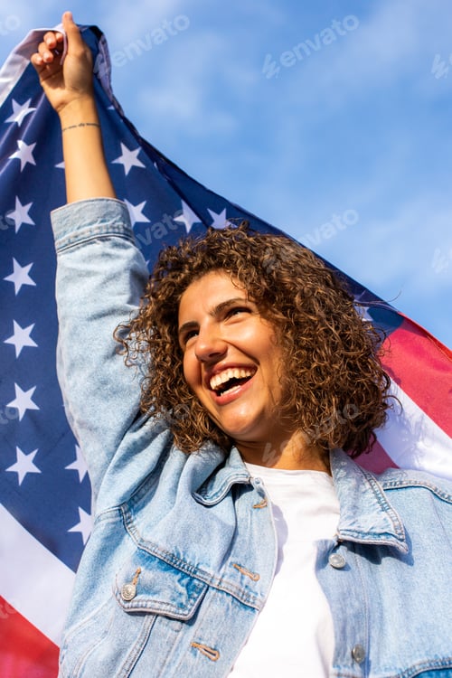 Preview: Happy Beautiful diverse woman holding United States of America flag and standing against blue sky