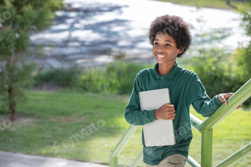Preview: A black curly haired African boy hugs his laptop and smiles at the camera at the railing under the s