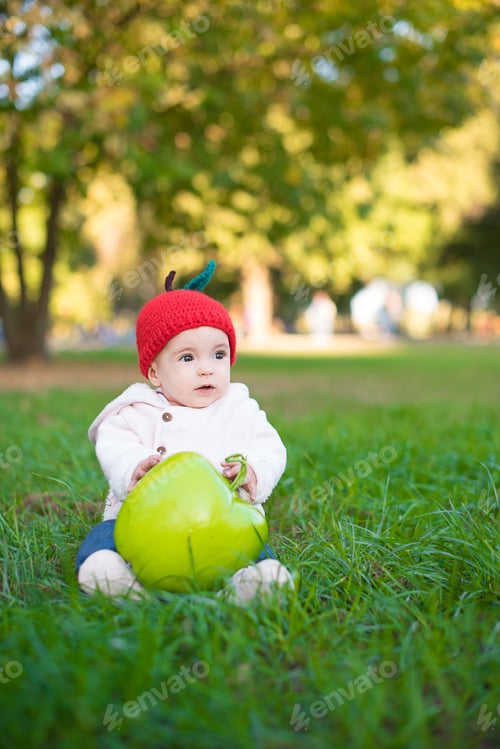 Preview: Cute baby girl in knitted hat sitting on the grass and holding the big apple.