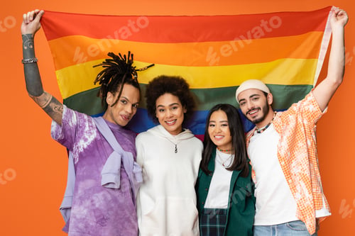 Preview: cheerful and trendy multiethnic friends posing with lgbt flag isolated on orange