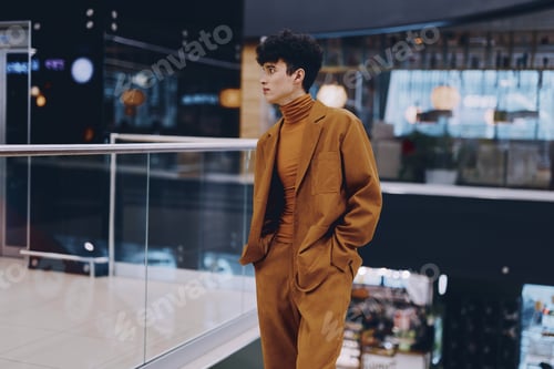 Preview: Stylish young man in a brown suit standing in a modern shopping mall with glass railings and ambient