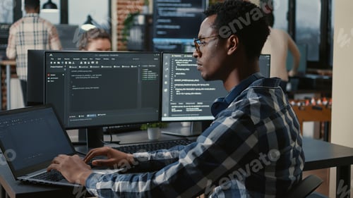 Preview: Focused programer fixing glasses while typing on laptop sitting at desk with multiple screens