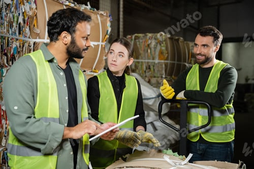 Preview: Smiling worker in reflective vest and gloves talking to multiethnic colleagues with digital tablet