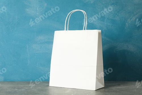 Preview: White paper bag on grey table against blue background, copy space