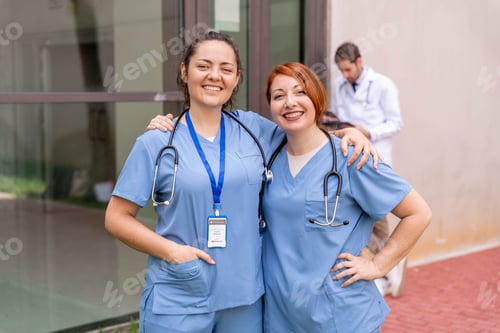 Preview: Two happy female nurses in scrubs smiling outside hospital building, colleague in background
