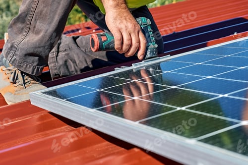 Preview: Man's hands with drill installing stand-alone photovoltaic solar panel system on rooftop of a house.