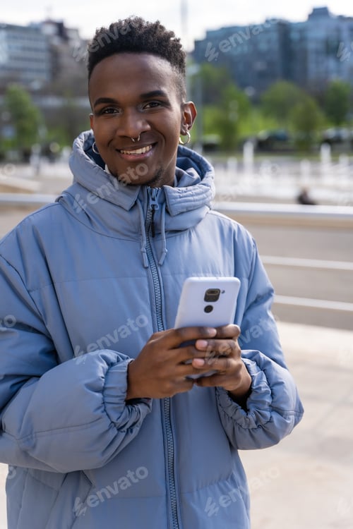 Preview: A black ethnic man with a phone and a blue jacket in the city, looking at social networks