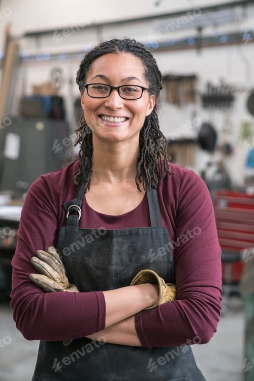 Preview: Woman in metal workshop, arms folded, smiling at camera.