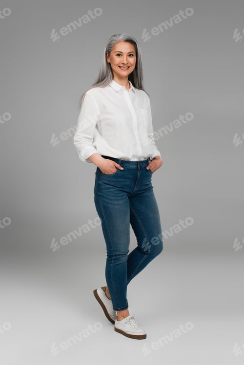 Preview: full length view of asian woman with hands in pockets of blue jeans smiling on grey