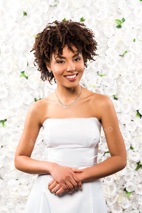 Preview: cheerful african american bride looking at camera while standing near flowers