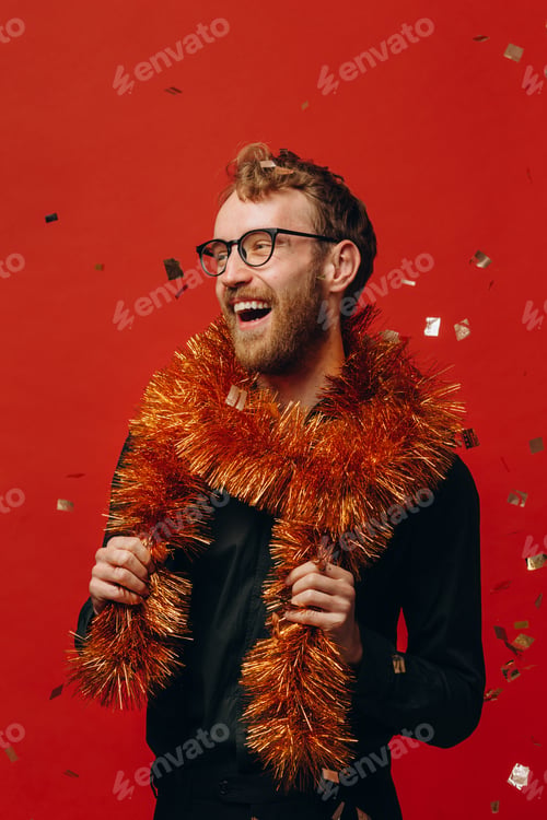 Preview: Red-haired cheerful man in glasses and golden boa at New Year party with confetti. Studio photo.