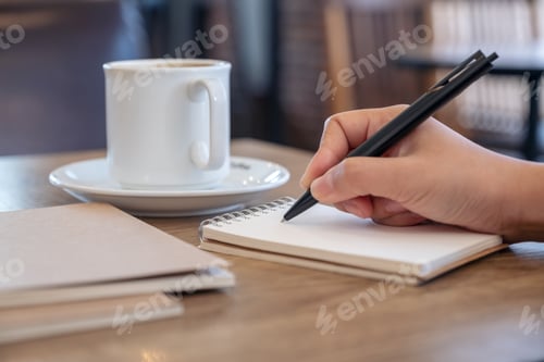 Preview: Closeup image of a hand writing on blank notebook with coffee cup on table in cafe
