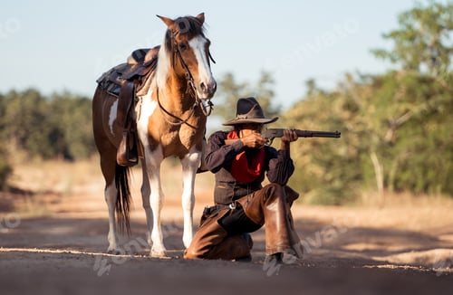 Preview: A cowboy kneeling on the ground while aiming a rifle beside his horse in a rugged outdoor setting