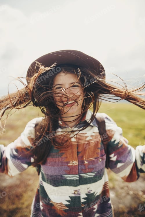 Preview: traveler hipster girl with windy hair and backpack, standing on top of sunny mountains