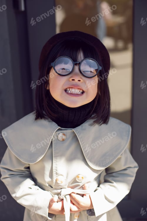 Preview: Korean girl in a gray coat and cap and round glasses stands the street in spring at the glass doors