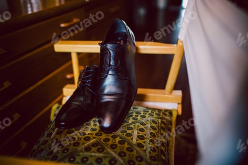 Preview: Black men's shoes, elegant and leather, resting on a chair on the bottom of a dark house.