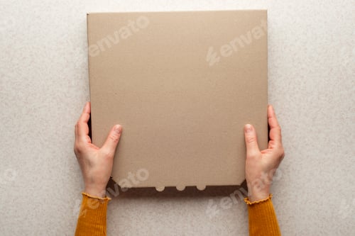 Preview: Pizza closed carton box in caucasian hands on kitchen table flat lay mockup with copy space