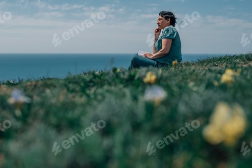 Preview: adult woman thoughtfully writes plans diary on a sunny day on a mountain against backdrop of the sea