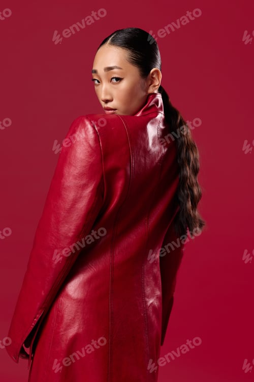 Preview: Fashion asian brunette in a red leather jacket posing against a red background with hands on hips