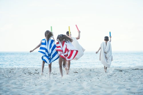 Preview: back view of children with towels as superhero robes and water toys in hands running on beach