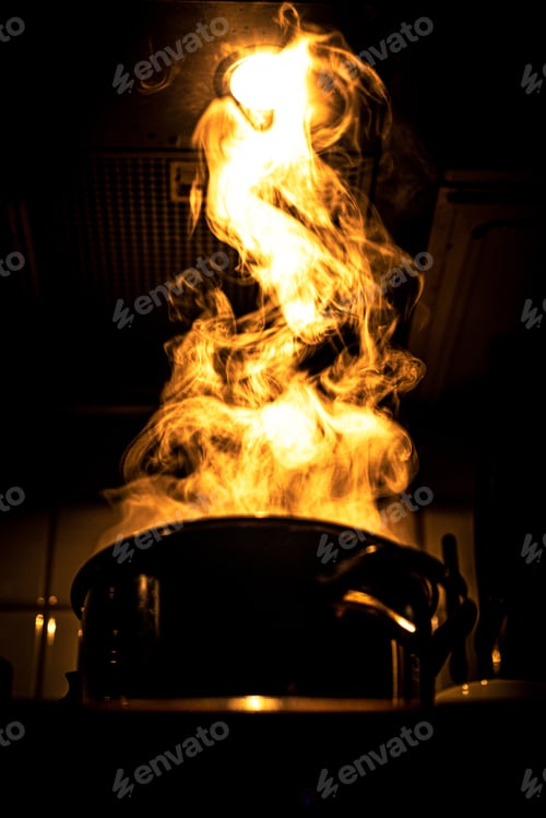 Preview: Vertical shot of a saucepan on fire with huge firery flames shooting up in the dark kitchen