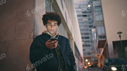 Preview: Cool hipster using smartphone at evening street in earphones closeup.