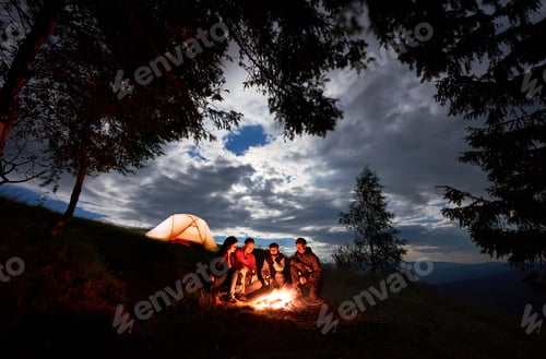 Preview: Four person are sitting around fire with beer near the camping and trees at dawn