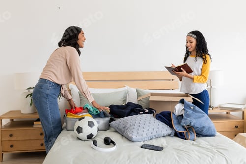 Preview: Mother and daughter organizing clothes and sports gear on bed, smiling together