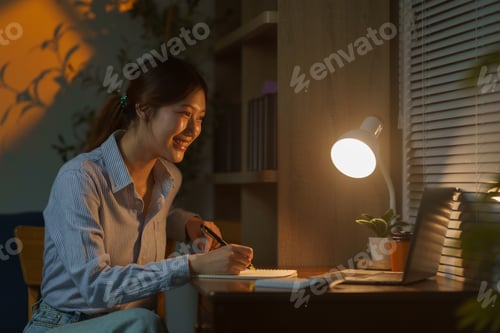 Preview: Young Asian Woman Working from Home at Night Taking Notes in Notebook and Smiling