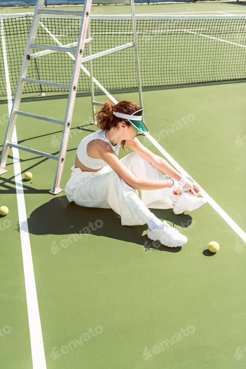 Preview: side view of young woman in stylish white clothing and cap tying shoelaces on tennis court with
