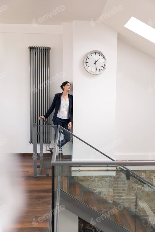 Preview: Businesswoman leaning against wall in office, waiting under clock