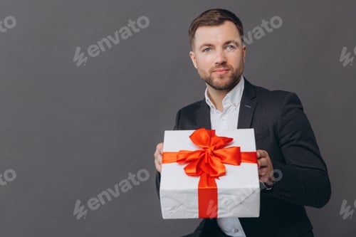 Vista previa: Hombre barbudo, feliz y sonriente en traje de negocios que sostiene un regalo y lo entrega sobre fondo gris.