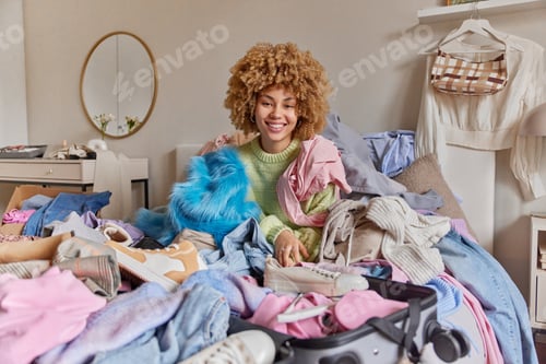 Preview: Indoor shot of cheerful curly haired woman packs things in suitcase prepares for travel poses in