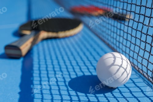 Preview: Ping pong ball by the net on blue table with paddles out of focus