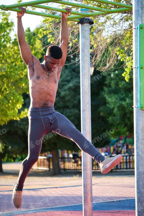Preview: Young fit shirtless black man exercising on monkey bars outdoors on sunny day.
