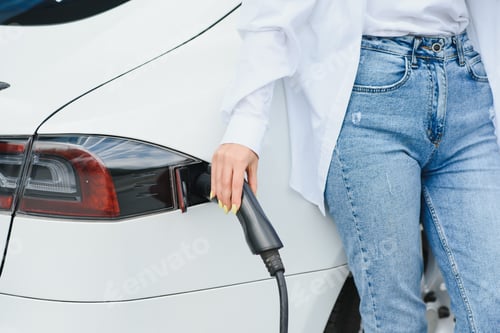 Preview: A girl's hand holds a cable for charging an electric car on the background of a white car.