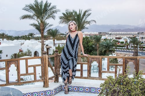 Preview: beautiful in dress looking at camera while leaning at wooden railing at resort in Egypt