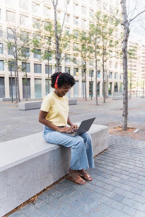 Preview: African American woman sitting on bench using laptop with headphones on campus